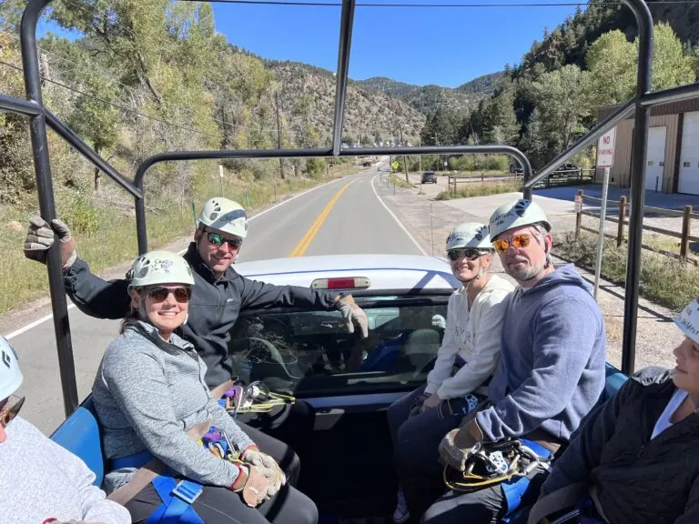 Group of five people in helmets and harnesses riding in an open-air vehicle on a mountain road with trees and hills in the background.