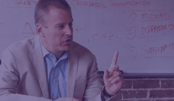 Man in beige blazer and blue shirt pointing at whiteboard with red handwritten notes about communication and risk.