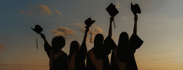 Four graduates raising their graduation caps in the air during sunset
