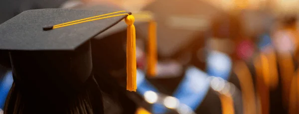Close-up of a graduation cap with a yellow tassel, blurred background of other graduates in caps and gowns.