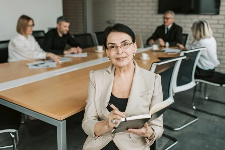 A businesswoman with short dark hair and glasses sitting at a conference table, holding a notebook and pen, with four colleagues in the background in a modern office setting.