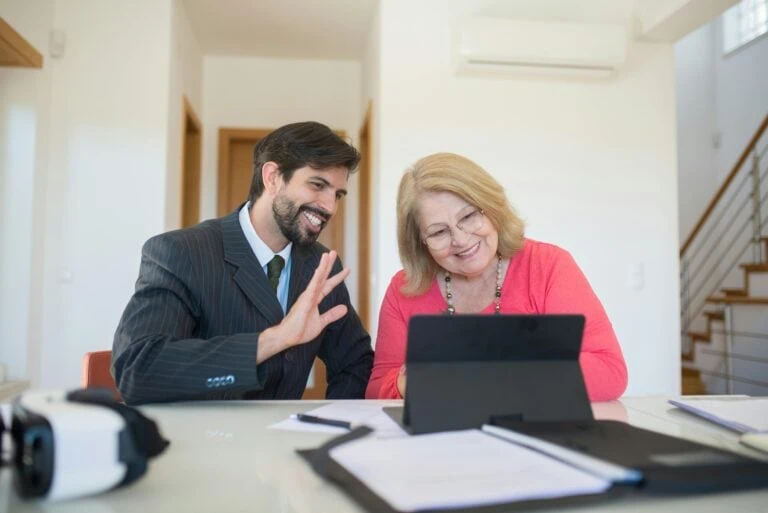 A man and an older woman smiling and waving at a laptop in a bright office space.
